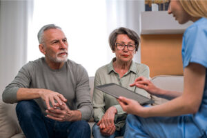 An elderly couple sits on the couch in their home while a nursing assistant in blue scrubs goes over their hospice plan on a tablet with them.