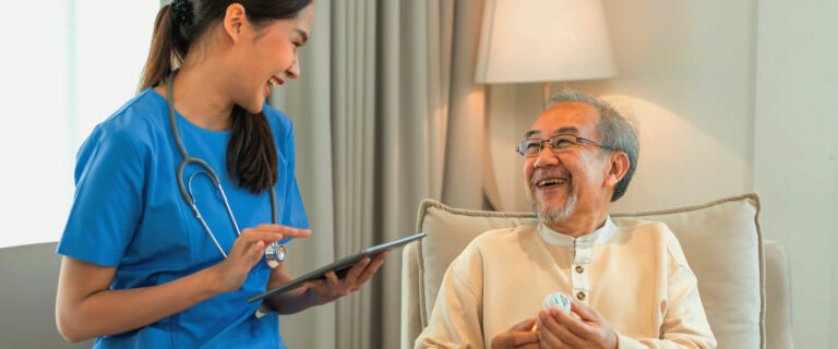 An elderly man sitting in a chair in his living room laughs together with his nursing aide.