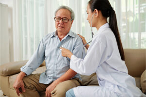 An elderly man in a button-down shirt sits on the couch in his living room while a doctor uses a stethoscope to listen to his heartbeat.