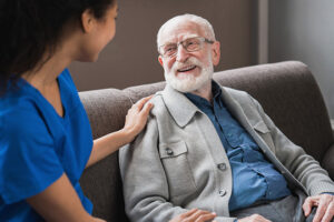 An elderly patient wearing glasses sits on his sofa in his living room while a care professional goes through memory care exercises with him.