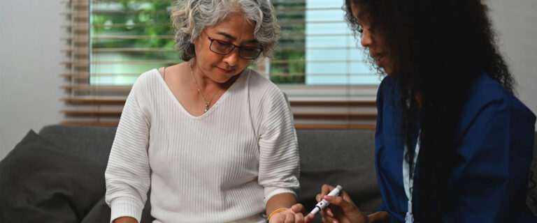 An elderly woman sitting on a couch in her home gets her finger pricked by a skilled nursing assistant to measure blood glucose levels.