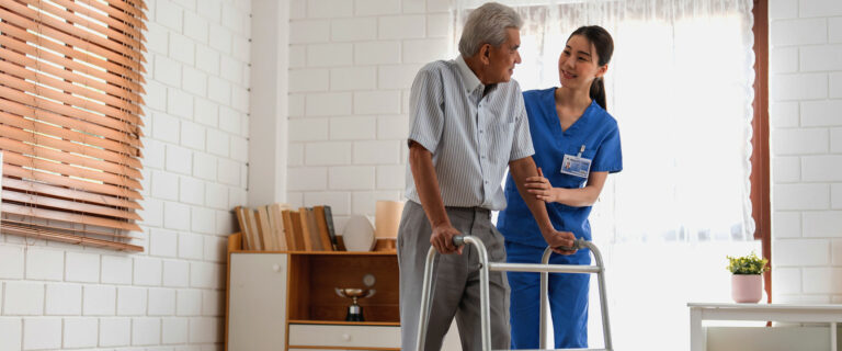 A Harmony Cares care professional in blue scrubs helps an older man using a walker move through his home.