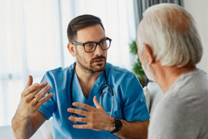 A male nurse in blue scrubs explains a fall risk prevention plan to an elderly male patient.