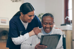 An elderly patient and her care professional laugh together while focusing on a tablet screen.