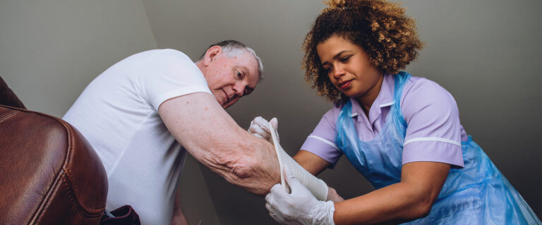 A home health aide helps an elderly man change the bandages on his wound.
